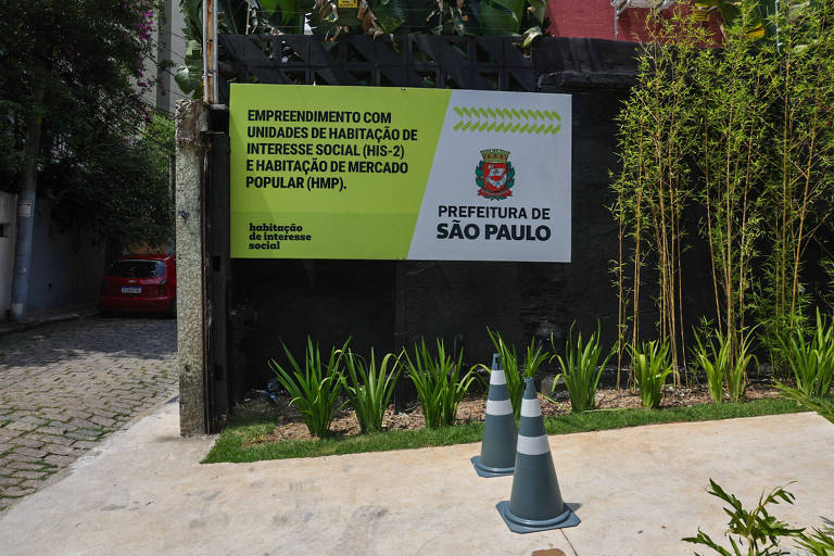 Placa verde e branca da Prefeitura de São Paulo anuncia empreendimento com unidades de habitação de interesse social e mercado popular, fixada em muro preto. Em frente, cones de sinalização e plantas ornamentais em área pavimentada e jardim com bambus ao lado.